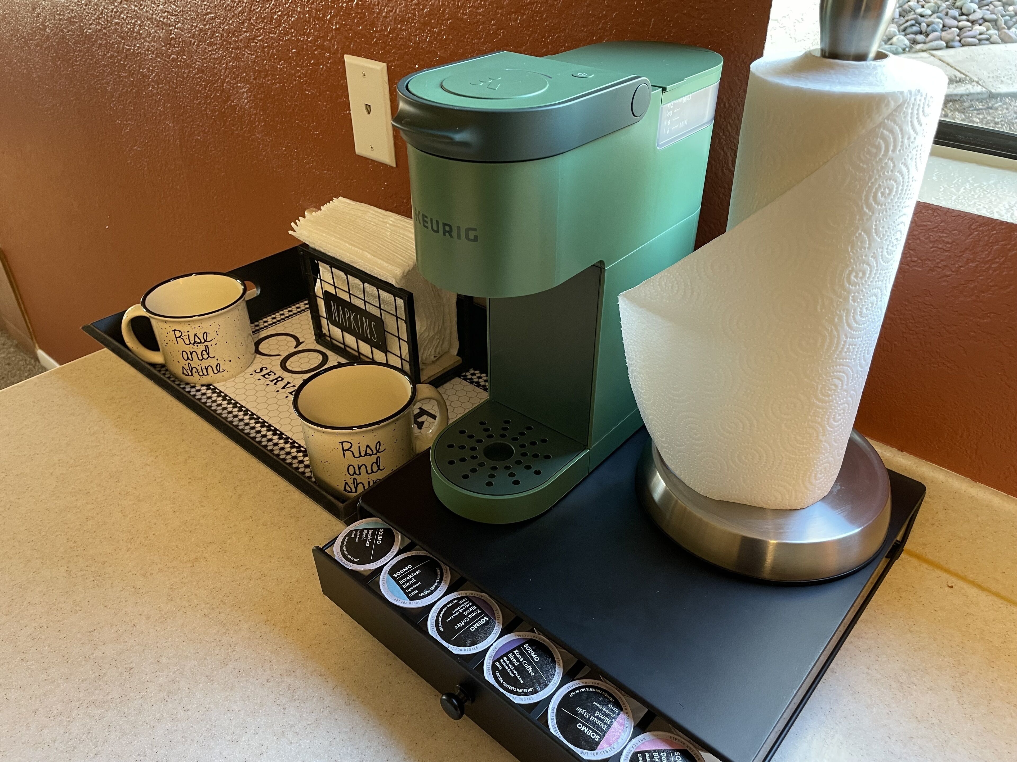 Uma cafeteira Keurig, um suporte para papel toalha e uma bandeja com cápsulas de café e canecas.