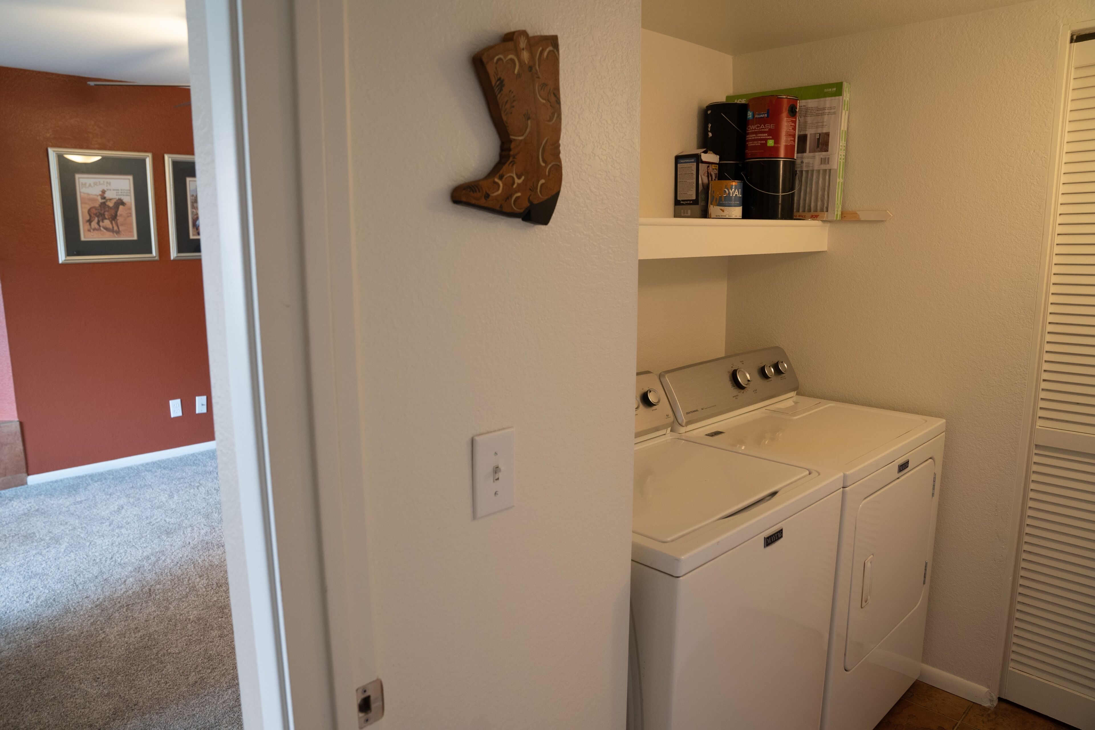 Un cuarto de lavado con lavadora y secadora, un estante con productos de limpieza y una puerta que da a otra habitación.