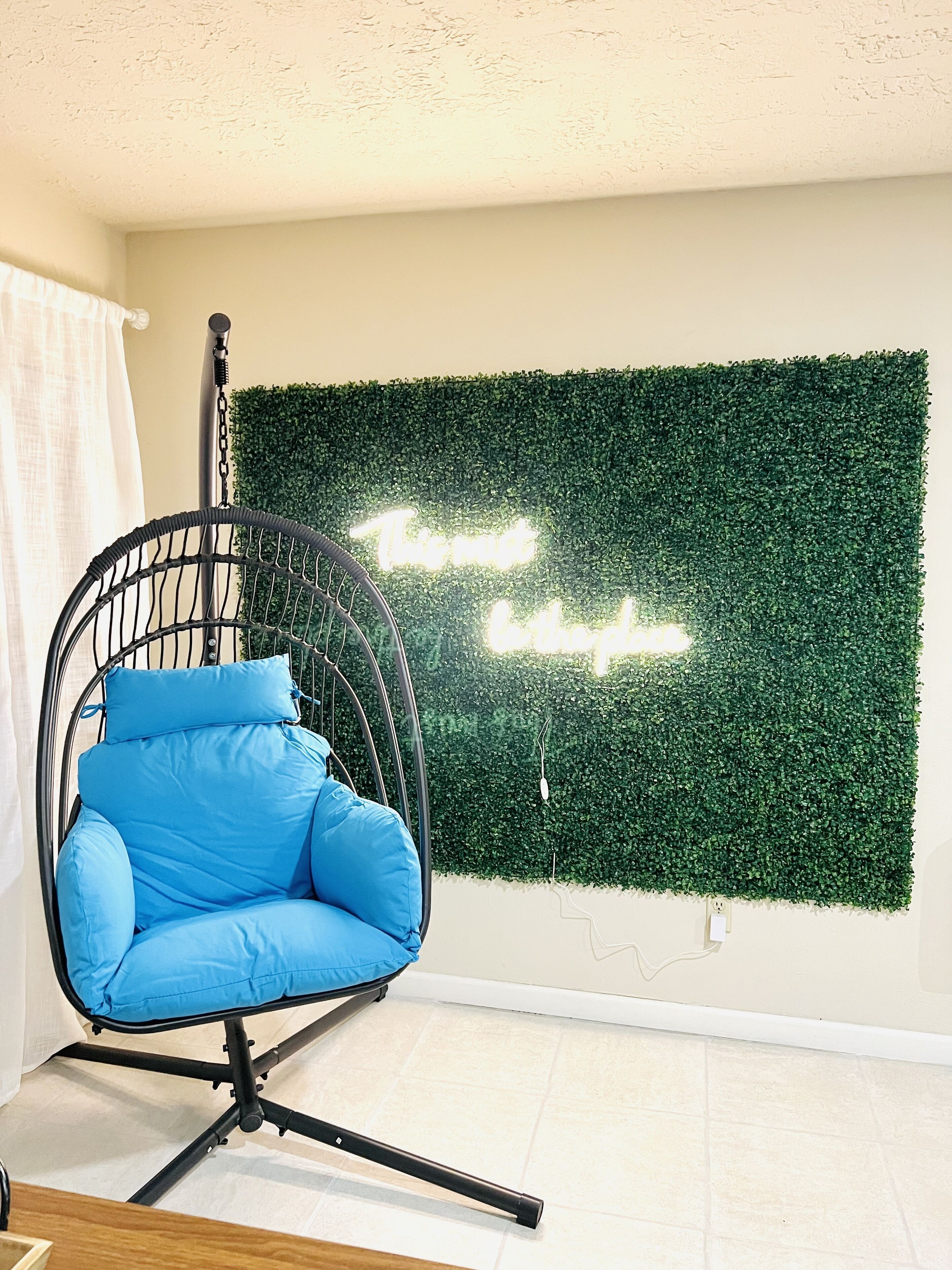 Een kamer met een hangstoel, een groene muur met een neonbord en een wit gordijn.