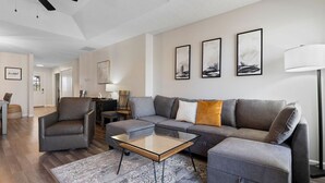 A modern living room with a sectional sofa, a glass coffee table, a leather armchair, and a floor lamp. There are abstract paintings on the wall and a ceiling fan. - Cozy Modern Branson Game House | 9 Min to SDC (Branson West)