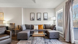 A modern living room with a sectional sofa, a glass coffee table, and a patterned rug. There are three framed artworks on the wall, a lamp, and a window with blinds. - Cozy Modern Branson Game House | 9 Min to SDC (Branson West)