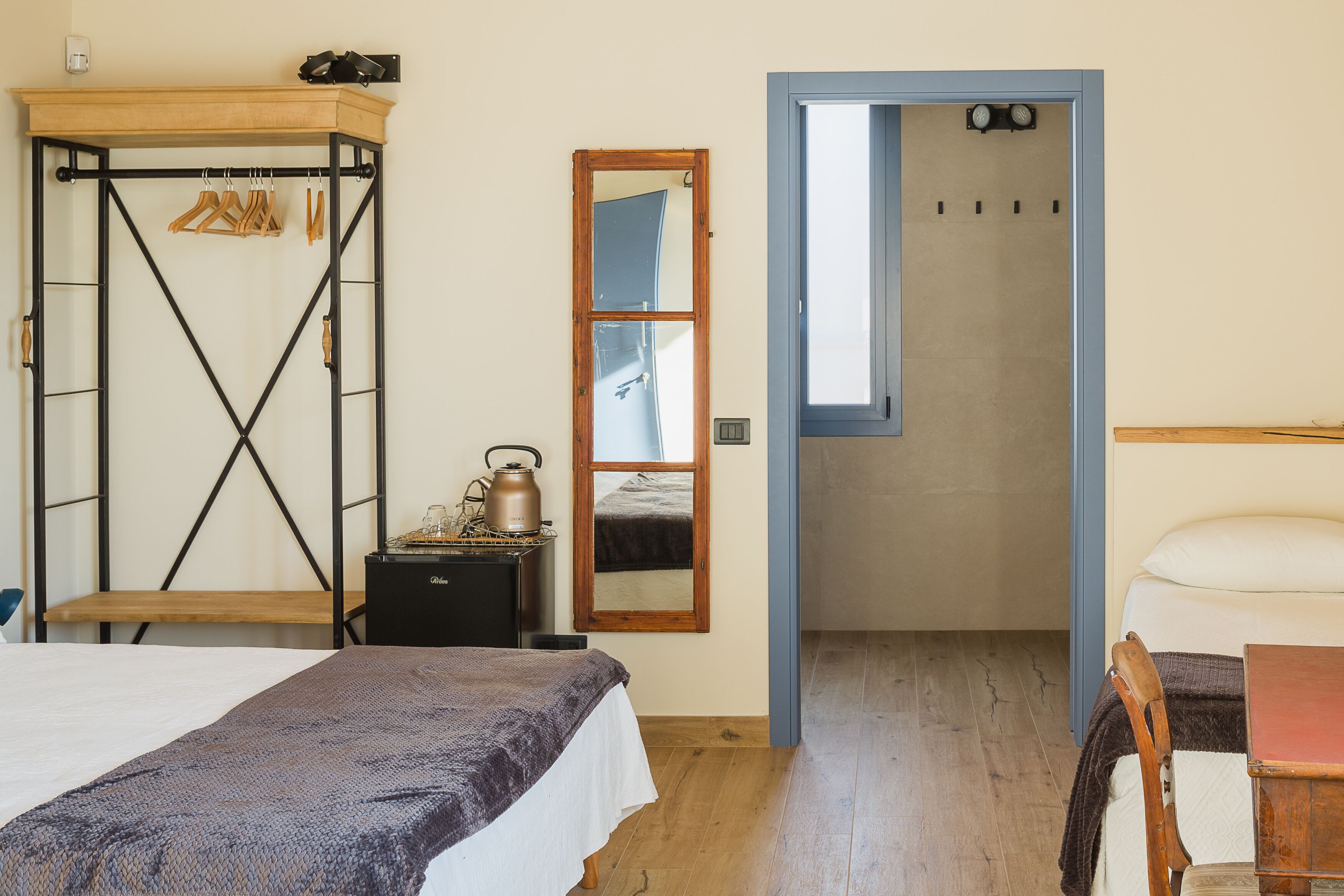 A room with a bed, a wooden bench, a small table with a kettle, a coat rack, and a mirror.