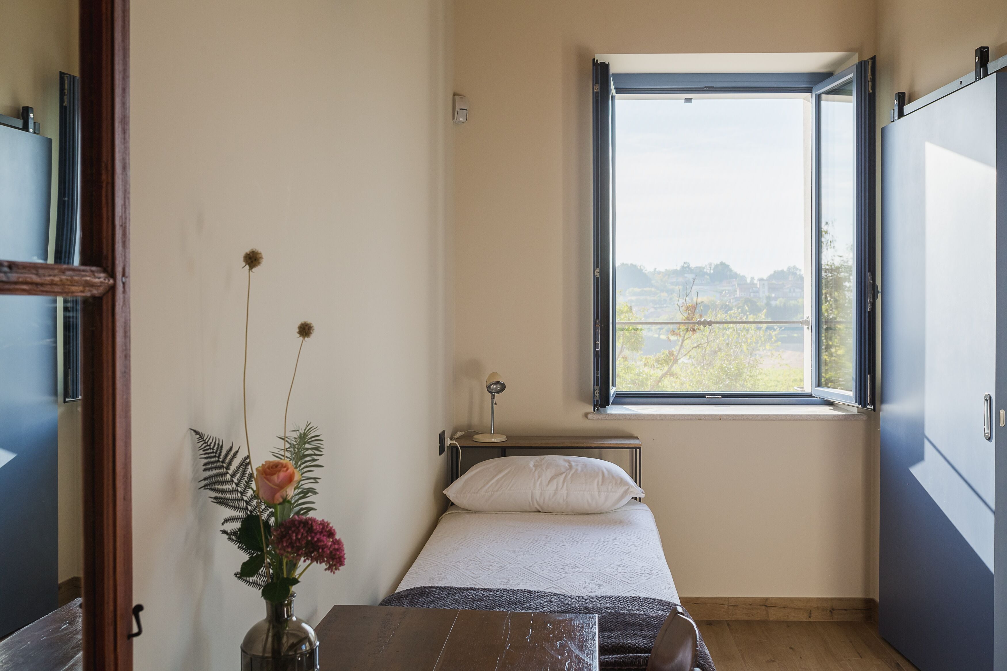 A bedroom with a bed, a window, a bedside table, and a vase with flowers.