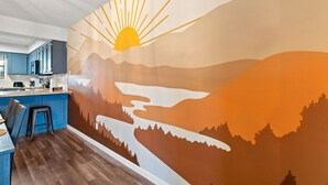 Keittiössä on siniset kaapit, baaritiski ja seinämaalaus, jossa on kuvattu auringonlasku maiseman yllä.