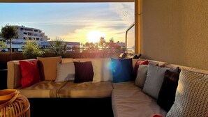 Een zonnig balkon met een bank en een rieten stoel, met uitzicht op een stadsbeeld bij zonsondergang.
