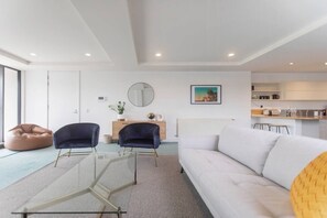 Una sala de estar moderna con un sofá, dos sillones, una mesa de centro de vidrio, un asiento de saco, una pequeña mesita de noche con espejo y una zona de cocina al fondo.