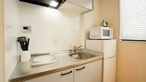 Una cocina con un lavamanos de acero inoxidable, un microondas y una tetera en la encimera. Hay un armario blanco debajo del lavamanos y una ventana con persianas a la derecha.