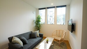 Una sala de estar moderna con un sofá gris, una mesa de centro de madera, un sillón de mimbre y un ventanal con vistas a edificios.