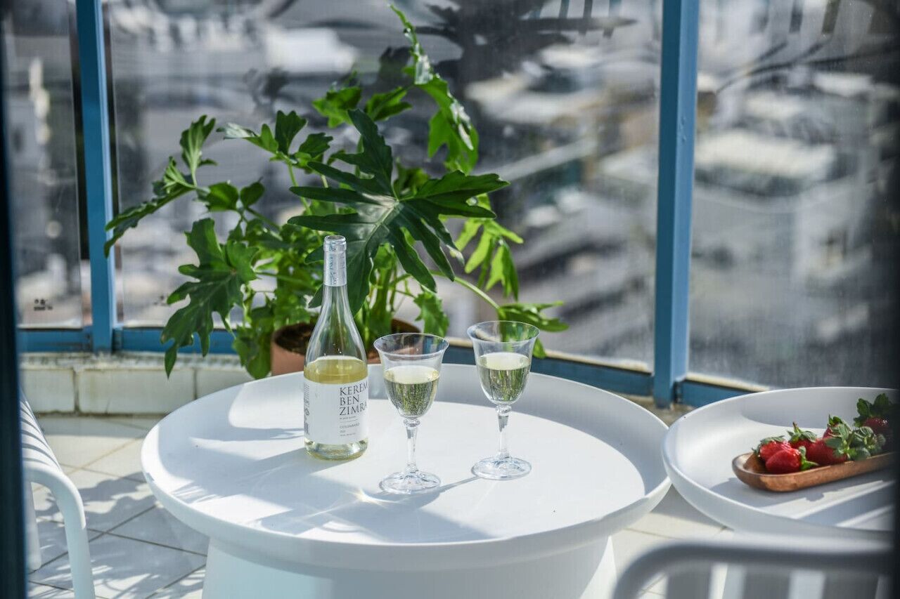 A table with a bottle and two glasses of white wine, a plate with strawberries, and a large green plant in the background.
