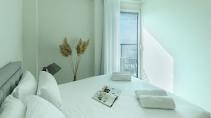 Habitación de hotel con una cama grande, ropa de cama blanca, una lámpara de noche, una revista y una ventana con vistas.