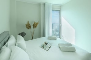Habitación de hotel con cama, ventana con cortinas, una revista sobre la cama y un jarrón con plantas secas.