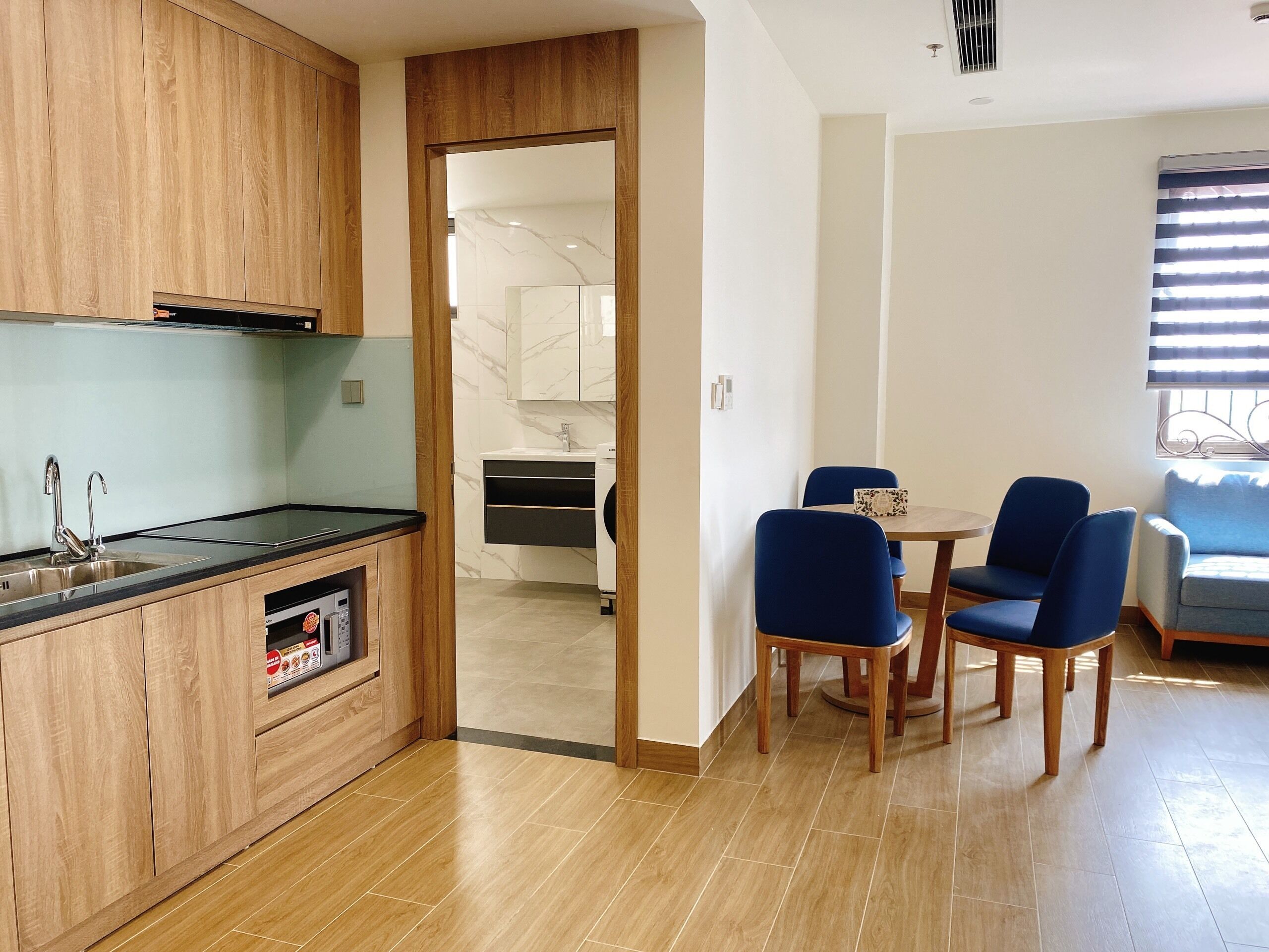 A modern kitchen with wooden cabinets, a built-in microwave, and a sink. There is a dining area with two blue chairs and a small table.
