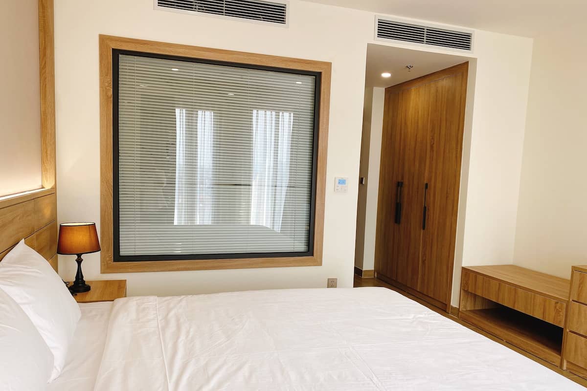 Ett hotellrum med en stor säng, en sänglampa, ett fönster med persienner, en garderob och ett litet sidobord.