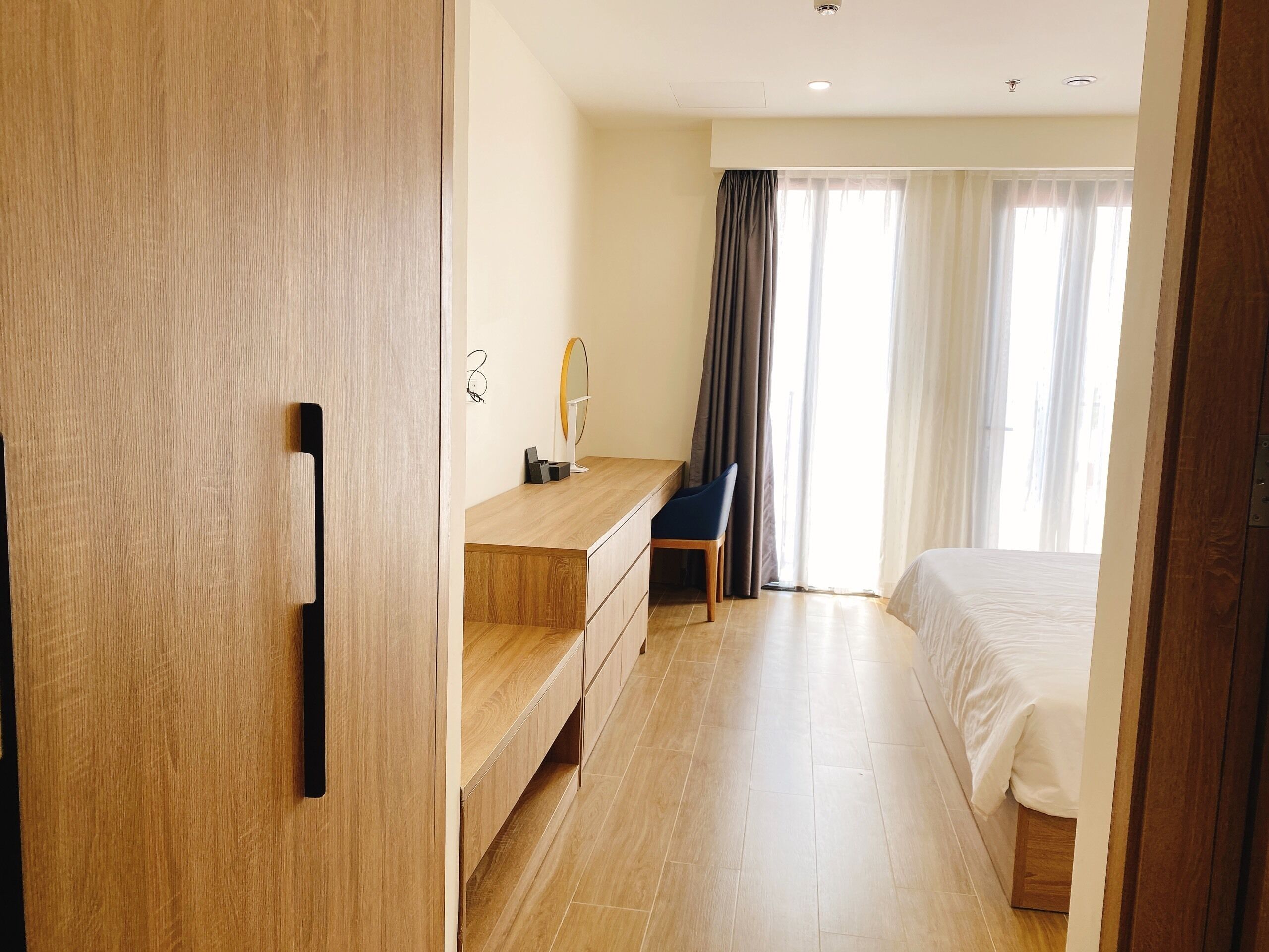 A modern hotel room with a wooden wardrobe, a bedside table with a mirror, a chair, and a bed.