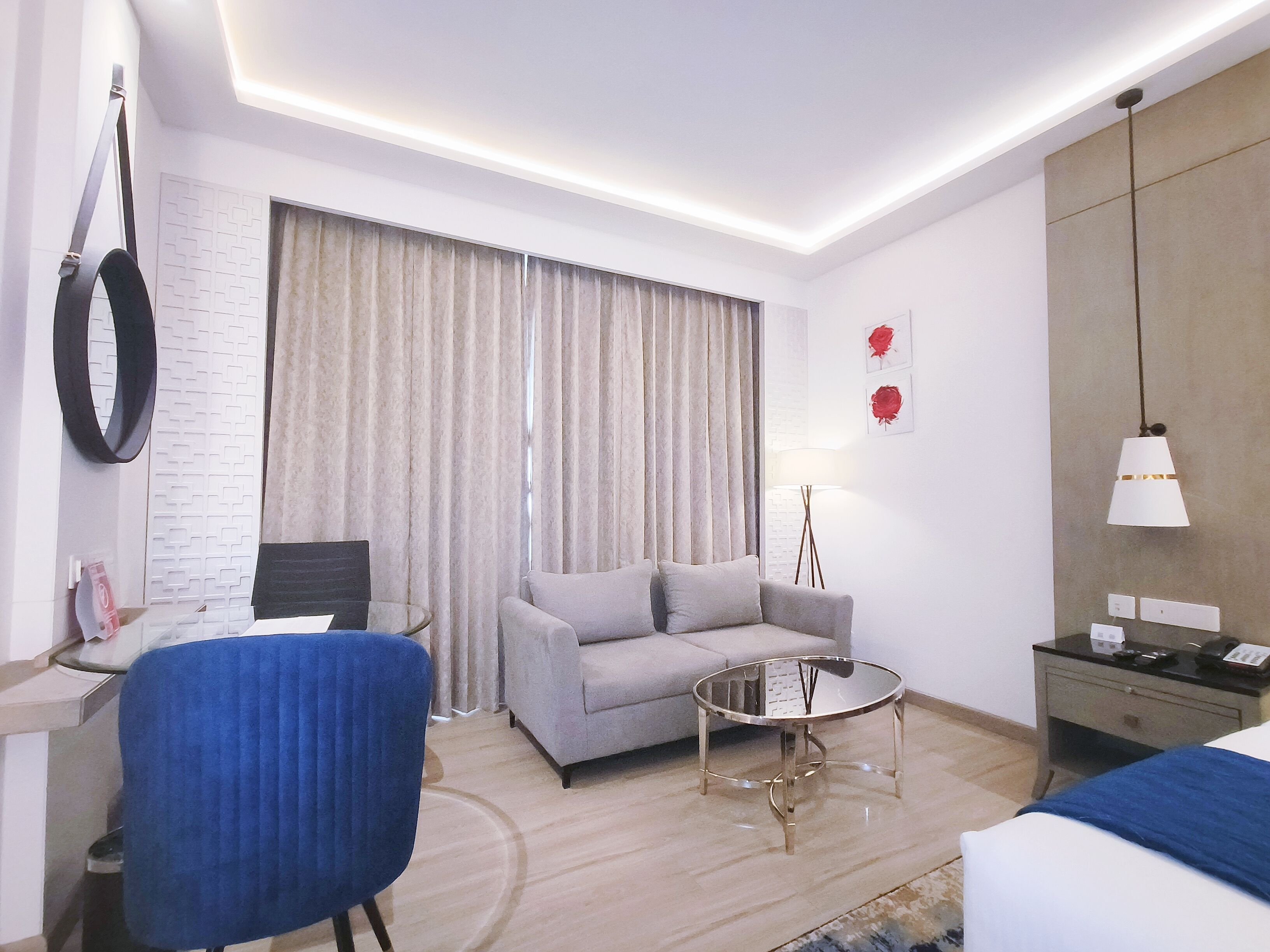 Una habitación de hotel moderna con un sofá gris, una mesa de centro redonda de cristal, un sillón azul, una cama con colcha azul, un escritorio con una computadora portátil, una lámpara y un espejo montado en la pared.
