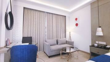 Una habitación de hotel moderna con un sofá gris, una mesa de centro redonda de cristal, un sillón azul, una cama con colcha azul, un escritorio con una computadora portátil, una lámpara y un espejo montado en la pared.