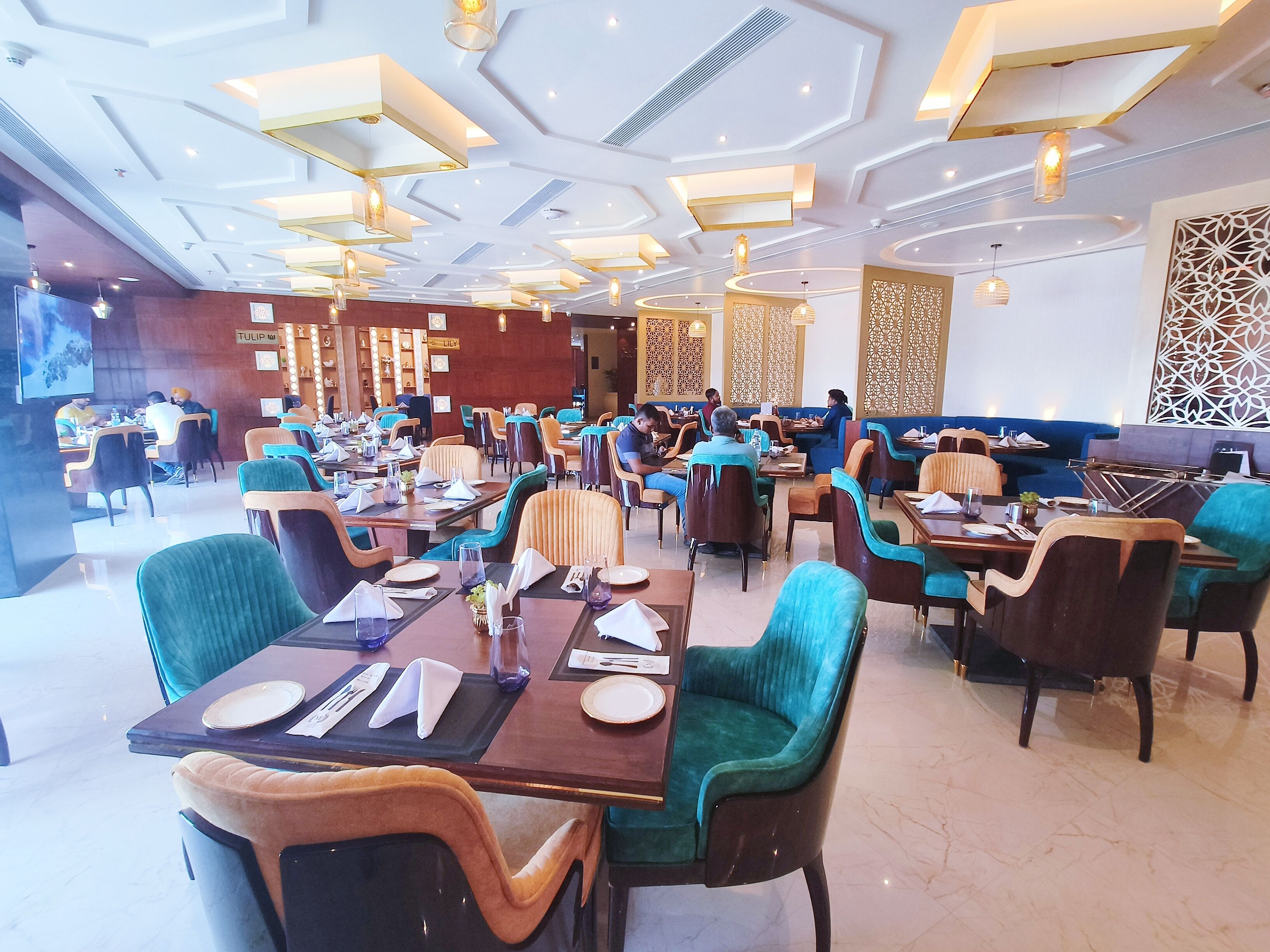 A modern restaurant with multiple dining areas, including a bar/lounge and a more formal dining section. Tables are set with plates, cutlery, and napkins. The ceiling features geometric patterns and recessed lighting.