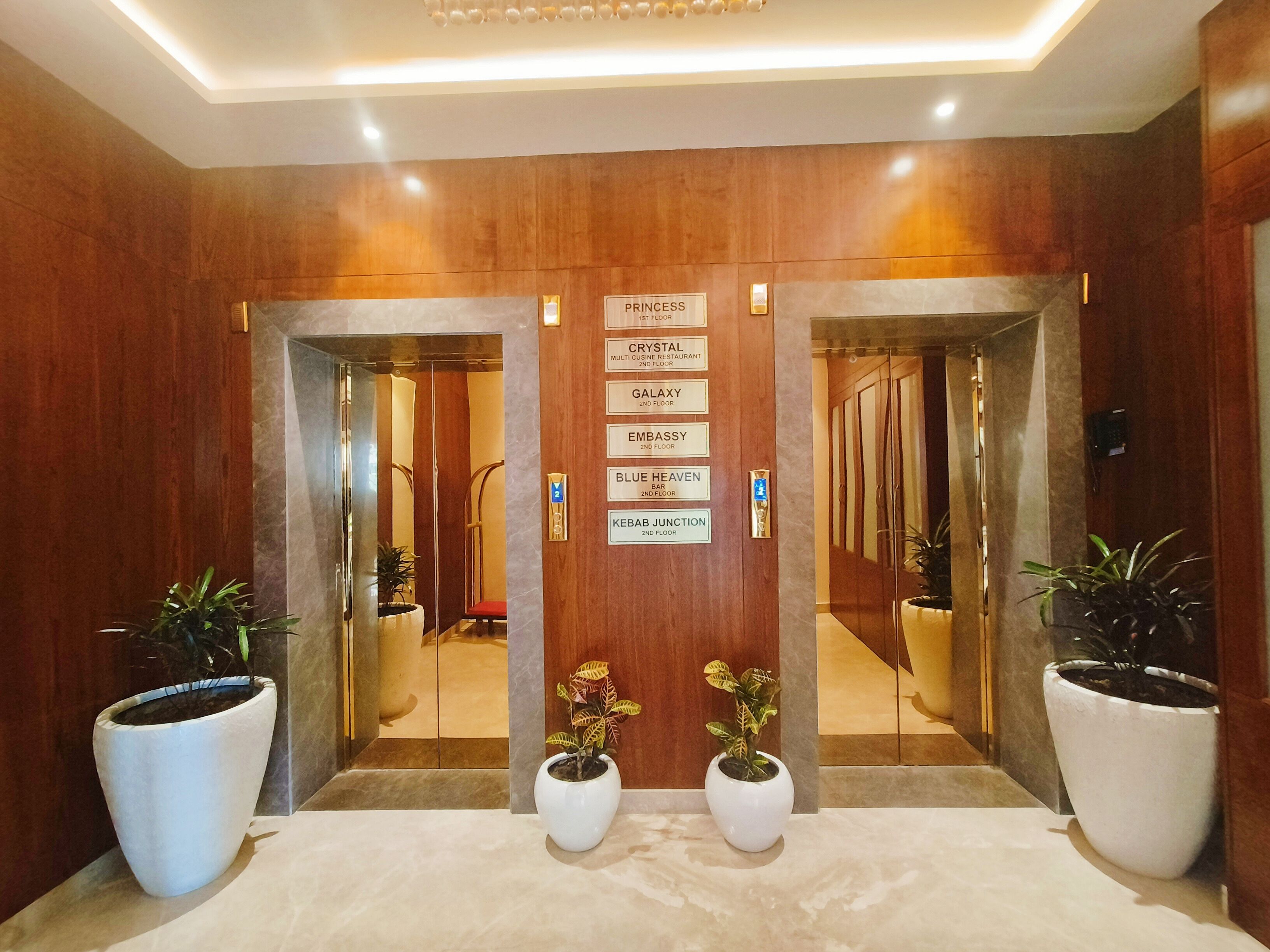 Un vestíbulo de hotel con un mostrador de recepción de madera, un cartel que indica la ubicación de Princess, Crystal, Galaxy, Embassy, Blue Heaven y Kebab Junction en el segundo piso.