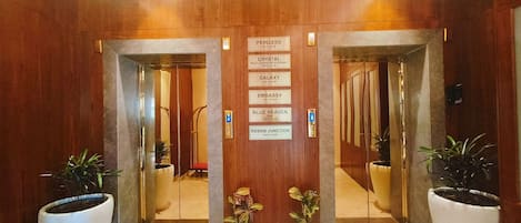 Un vestíbulo de hotel con un mostrador de recepción de madera, un cartel que indica la ubicación de Princess, Crystal, Galaxy, Embassy, Blue Heaven y Kebab Junction en el segundo piso.