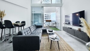 A modern living room with a grey sectional sofa, a dining area with black chairs, a flat-screen TV, and a large window offering a view of a building. - MAMAD - Luxe Apt in Top Tel Aviv Neighborhood (Tel Aviv)