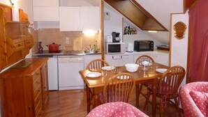 A compact kitchen with white cabinets, a wooden dining table, and chairs. There is a microwave, toaster, and a red pot on the counter.