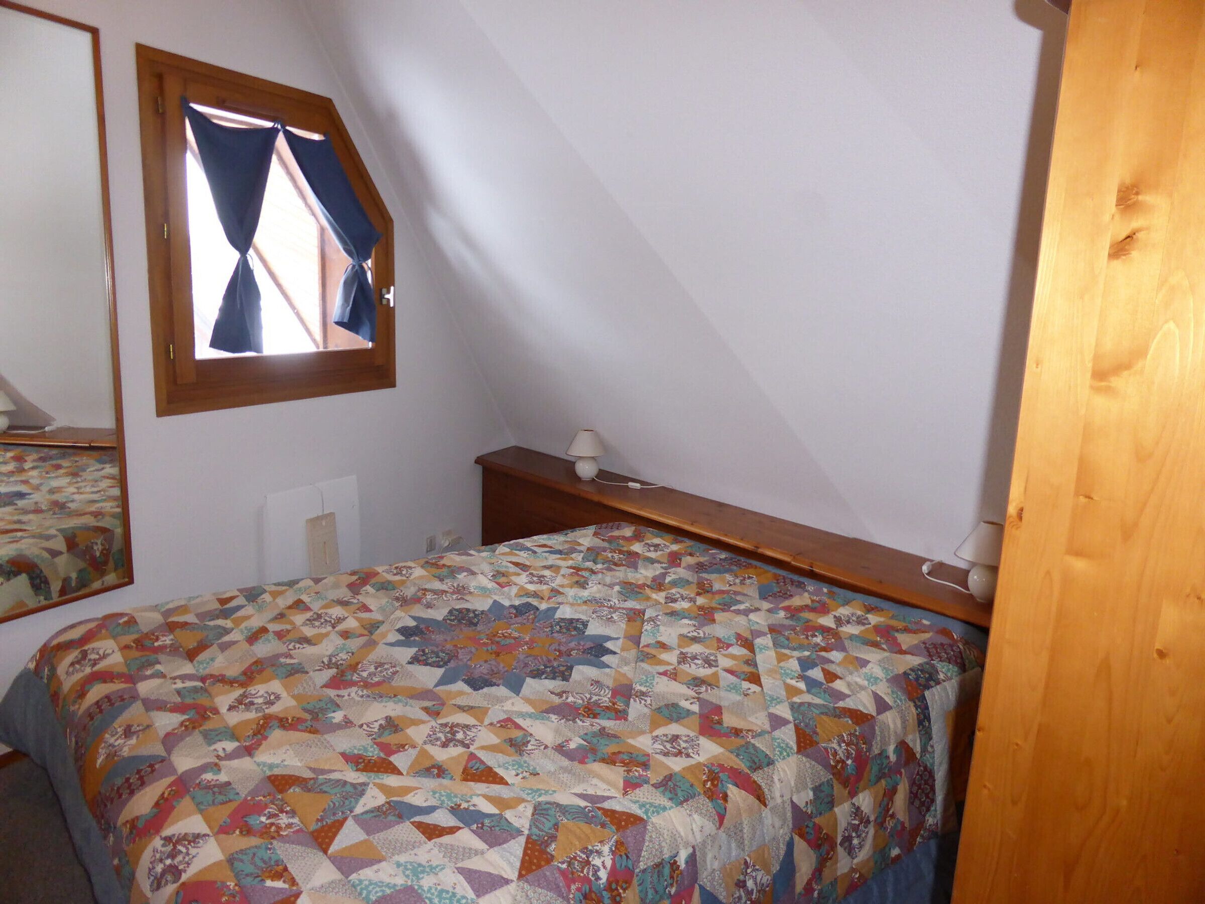 A bedroom with a sloped ceiling, a bed with a colorful quilt, a bedside table with a lamp, and a window with curtains.