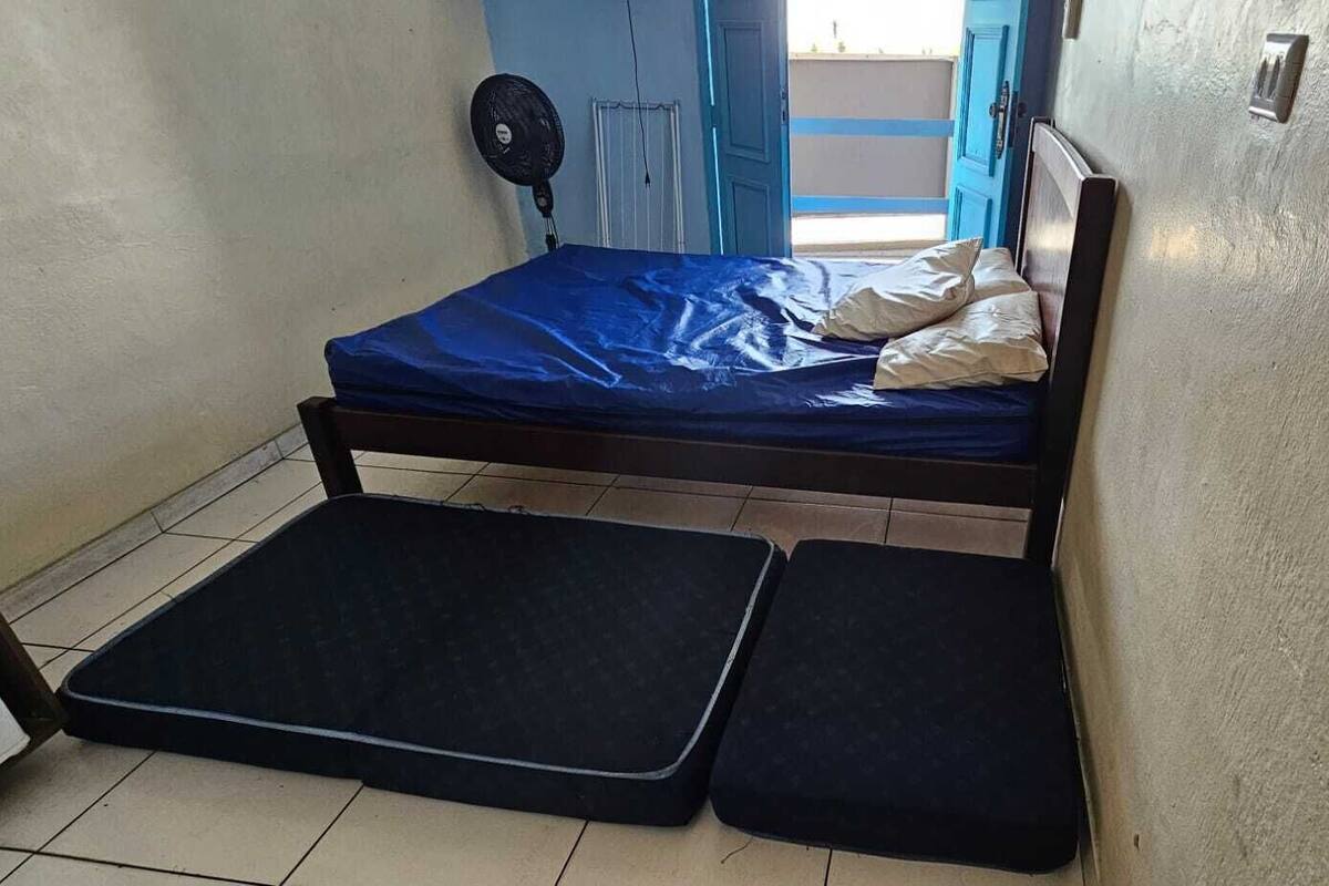 Une petite chambre avec un lit simple, un matelas bleu, une tête de lit en bois et un ventilateur fixé au mur.