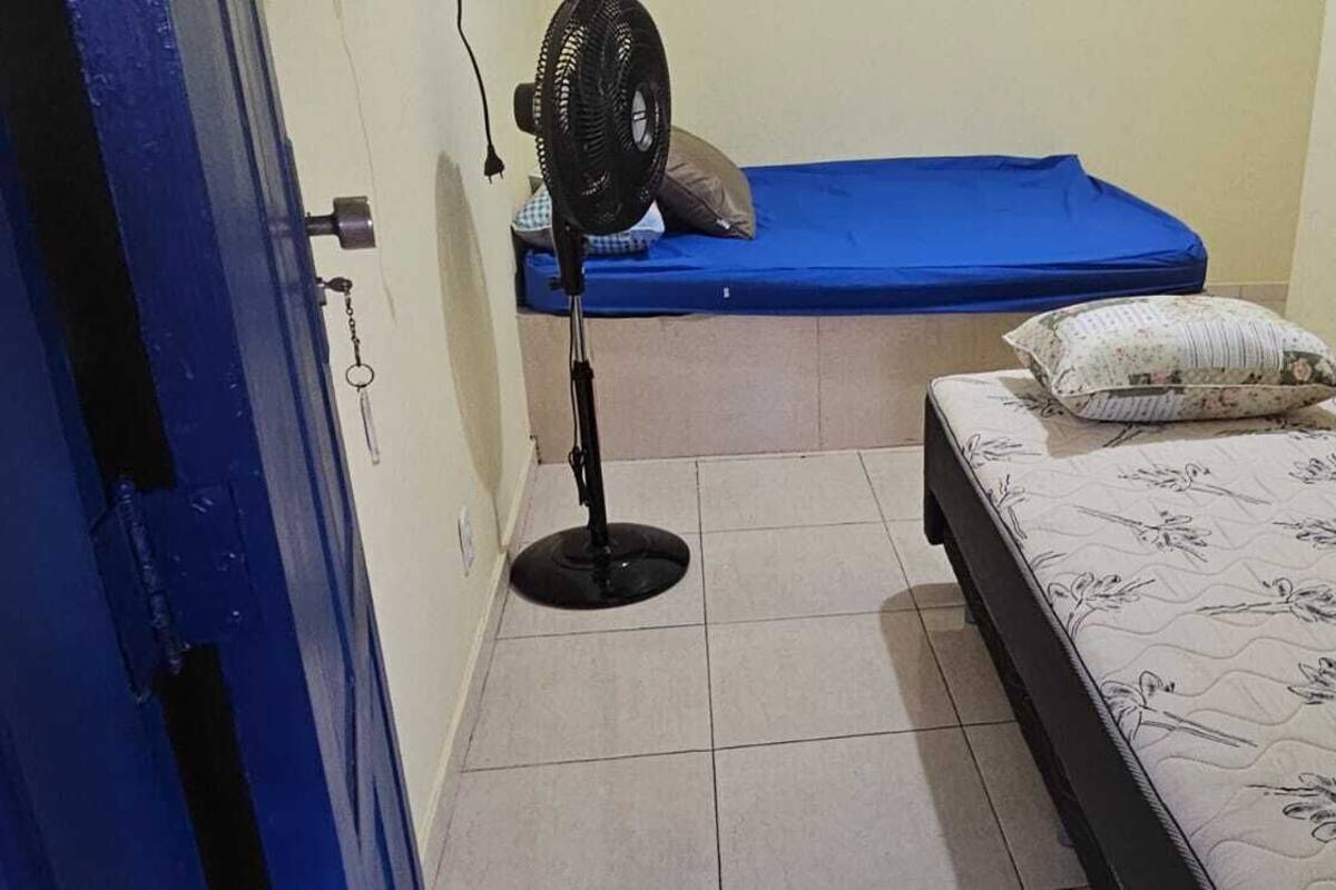 Une petite pièce étroite, avec un lit, un ventilateur sur pied et un matelas bleu.