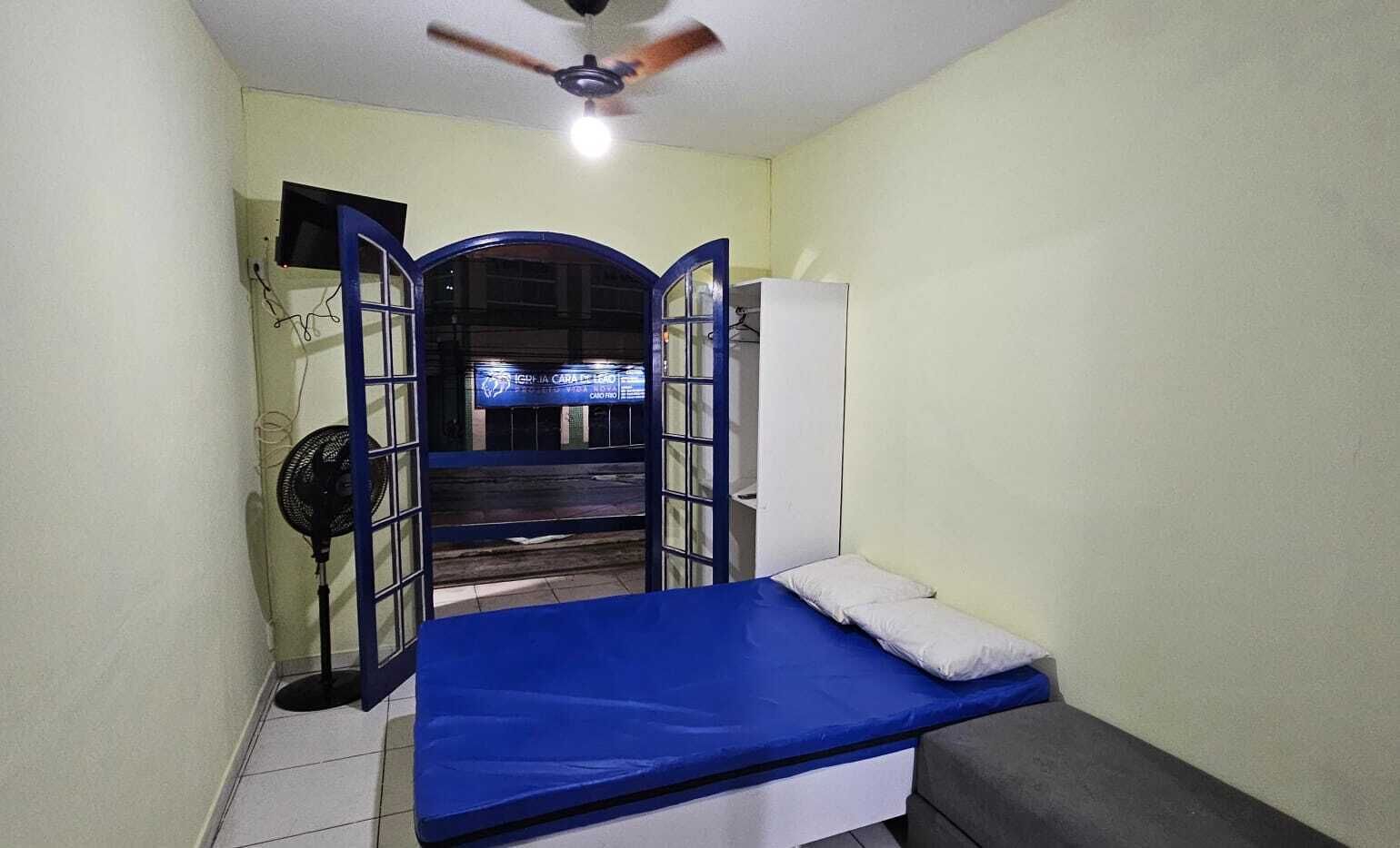 A small, narrow room with a single bed, a ceiling fan, a TV, and a refrigerator.