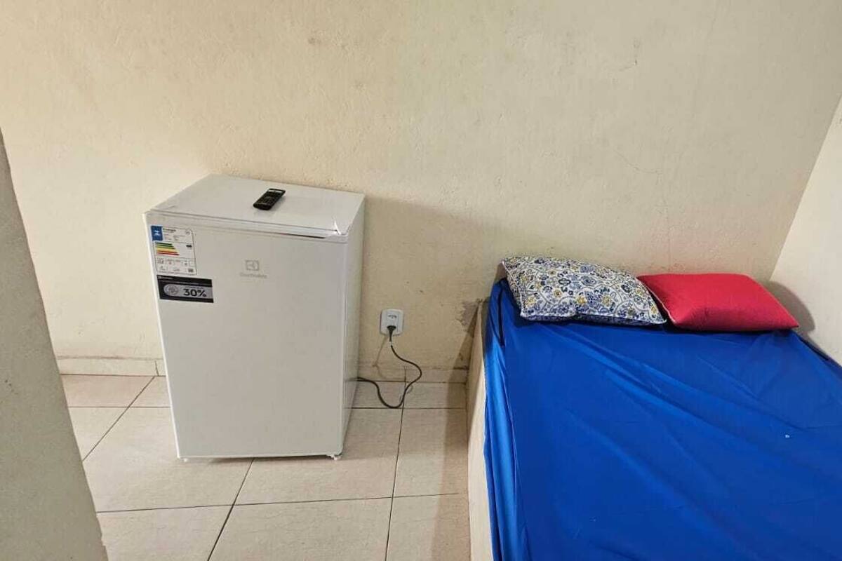 Une petite pièce avec un lit, un réfrigérateur blanc et une prise électrique au mur.