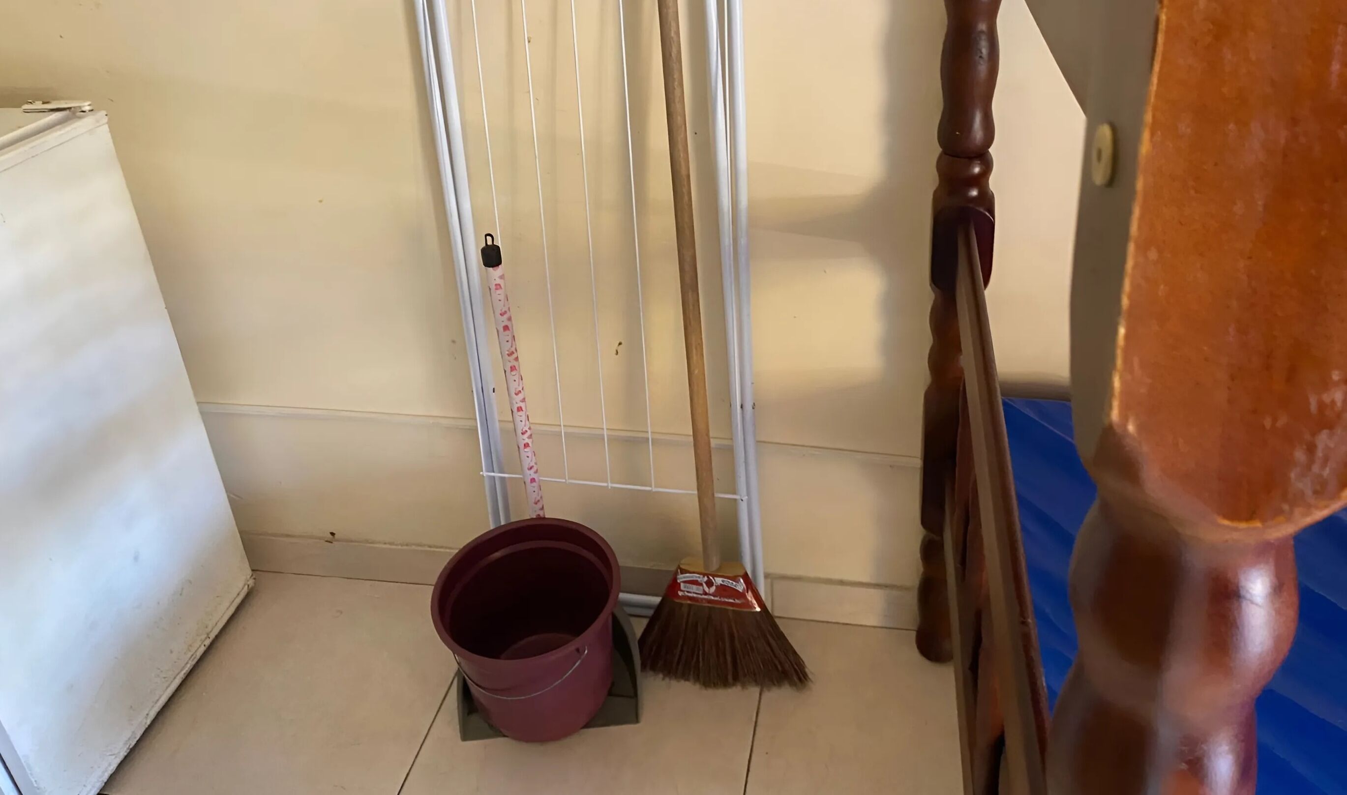 A corner of a room with a white radiator, a broom leaning against a white wall, and a red bucket on the floor.