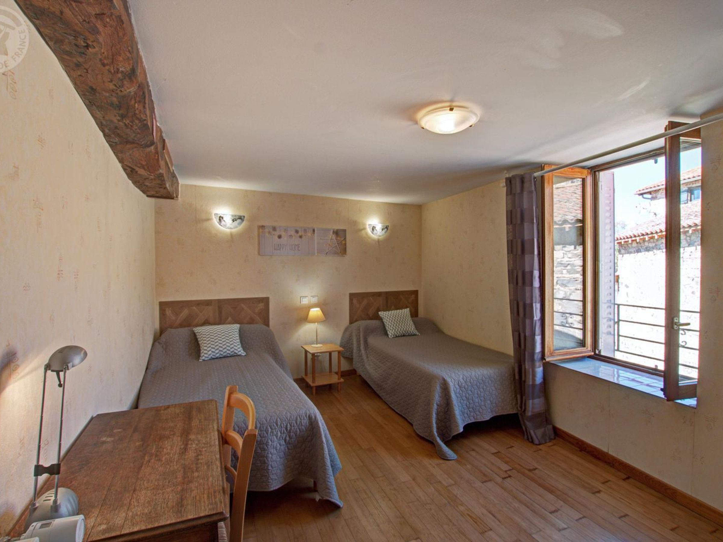 A room with two beds, a wooden table, a lamp, and a window with curtains.