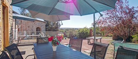 En udendørs terrasse med et bord og stole, en stor parasol og en bygning af sten i baggrunden.