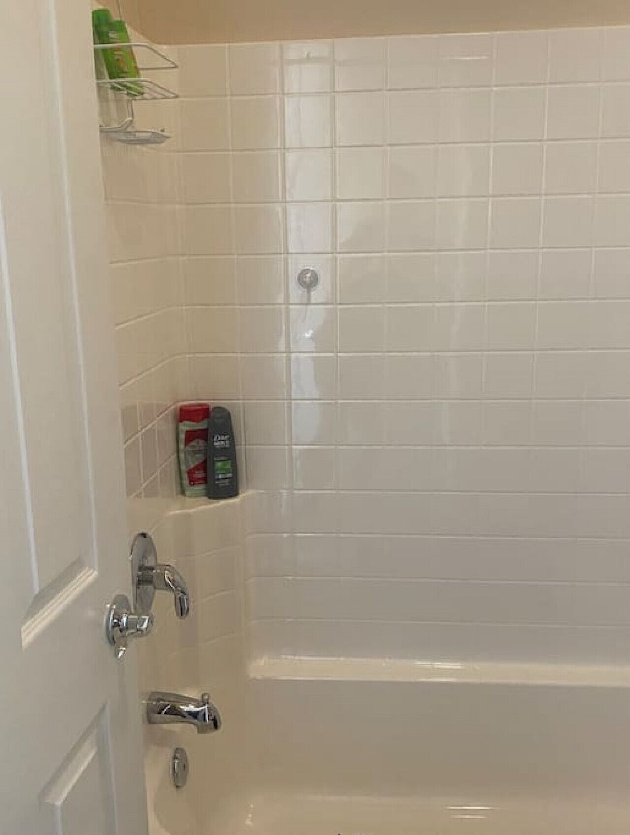 Une salle de bain avec des murs carrelés de blanc, une baignoire et une douche. On trouve des articles de toilette sur une étagère.