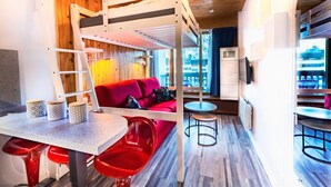 Habitación de hotel pequeña con una cama nido, un sofá rojo, una mesa de comedor y una zona de bar.