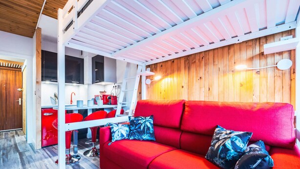 Un espacio habitable compacto con un sofá rojo, techo de madera y una zona de cocina con electrodomésticos rojos.