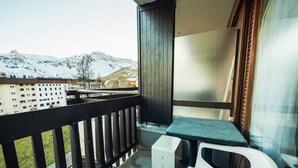 Un balcón con vistas a montañas nevadas, una mesita pequeña, una silla blanca y una ventana con cortinas.