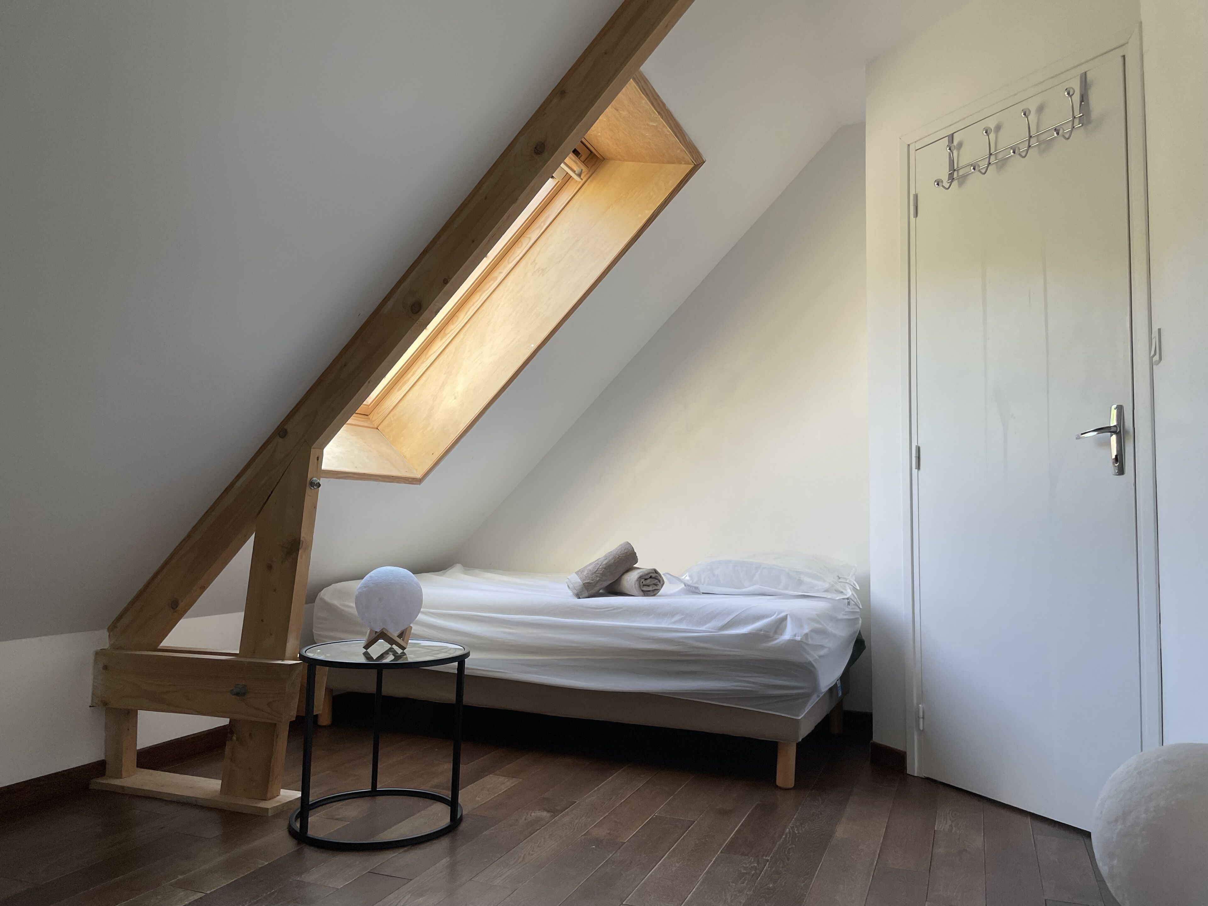 Una habitación pequeña con techo inclinado, una cama individual, una mesita redonda y una puerta con ganchos.