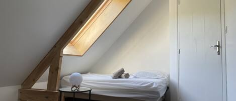 Una habitación pequeña con techo inclinado, una cama individual, una mesita redonda y una puerta con ganchos.