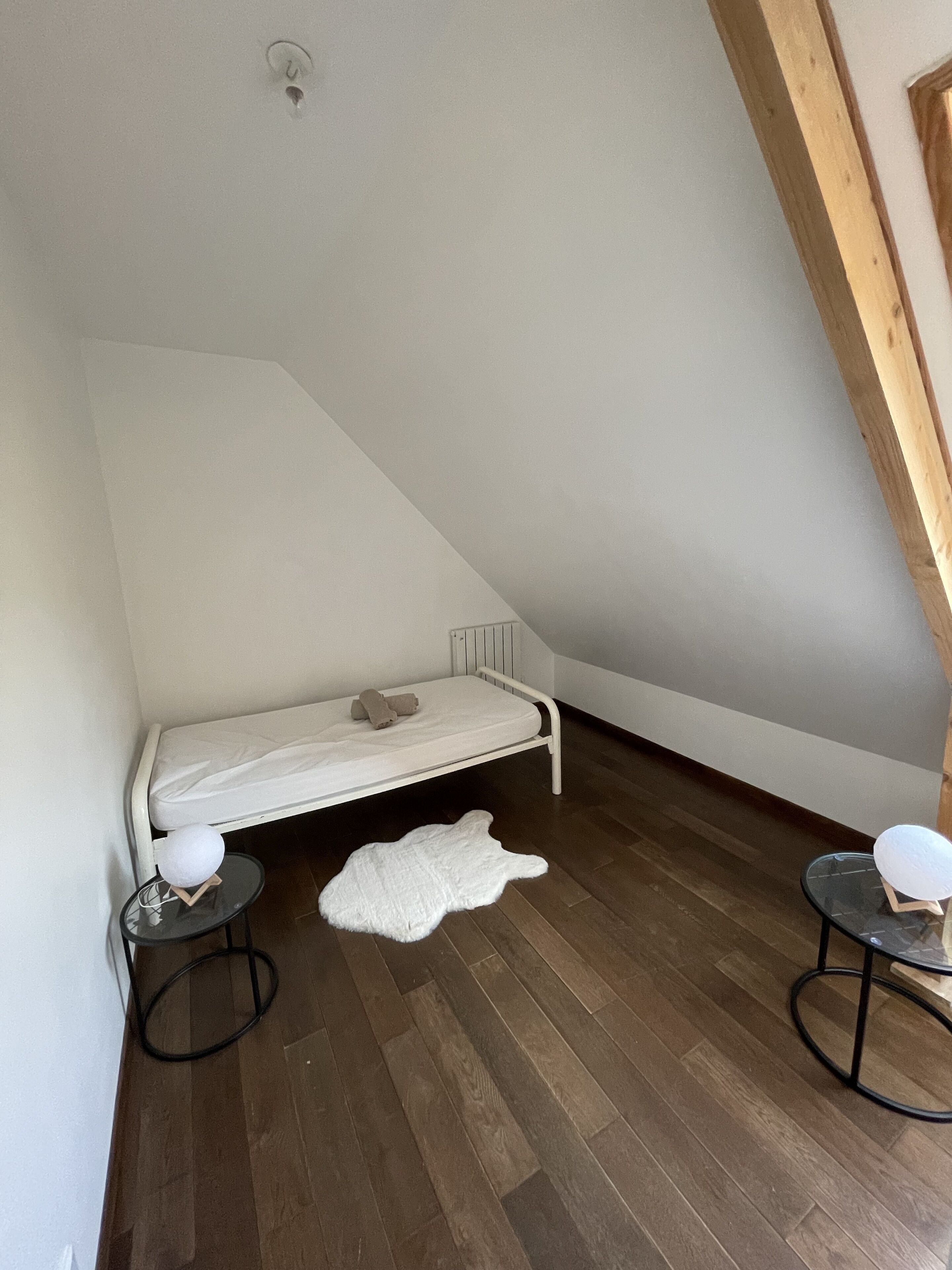 A small, slanted room with a single bed, two small tables, and a white rug.