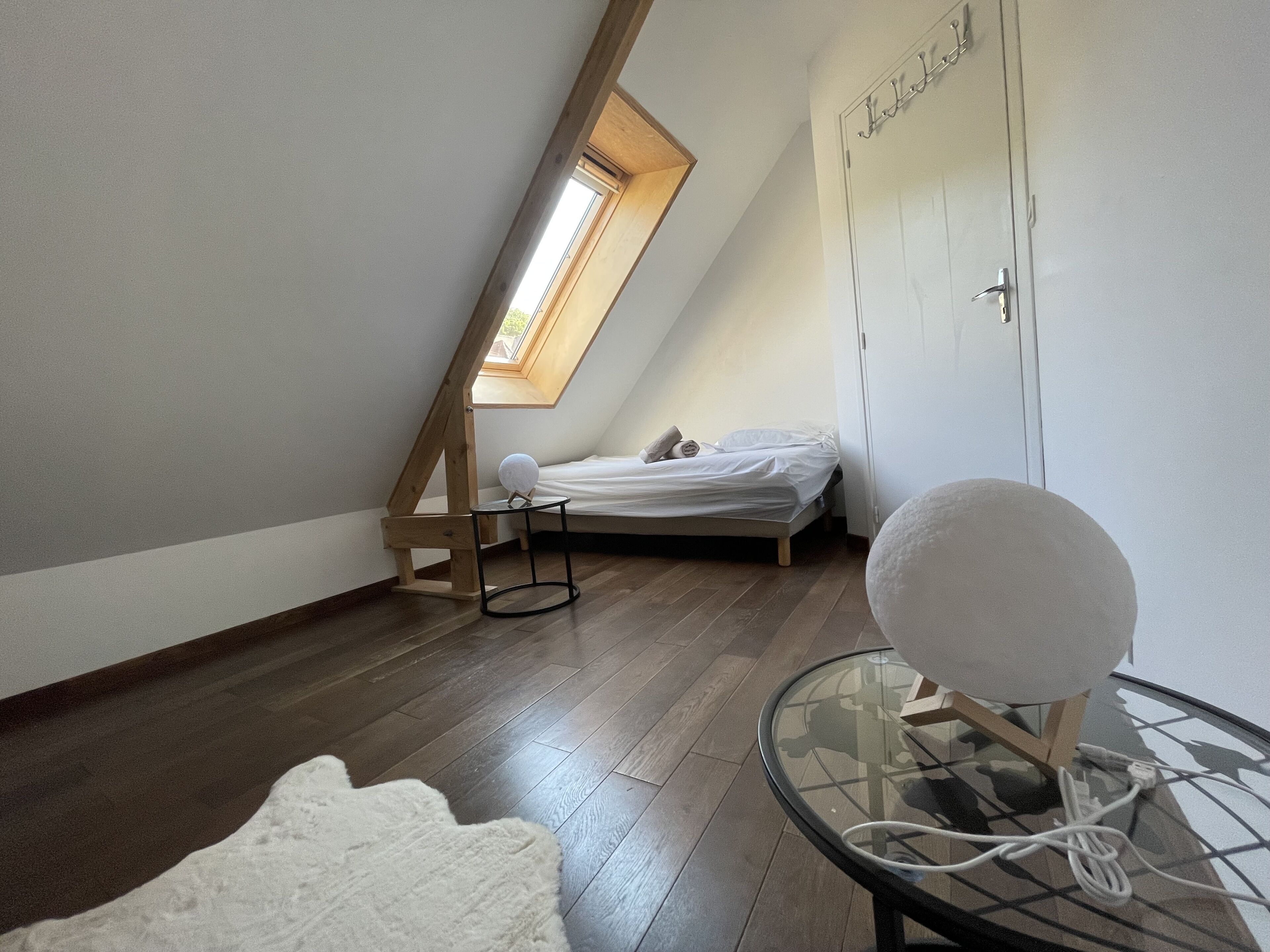A small, tidy room with a sloped ceiling, a bed, a round table with a glass top, and a white rug.