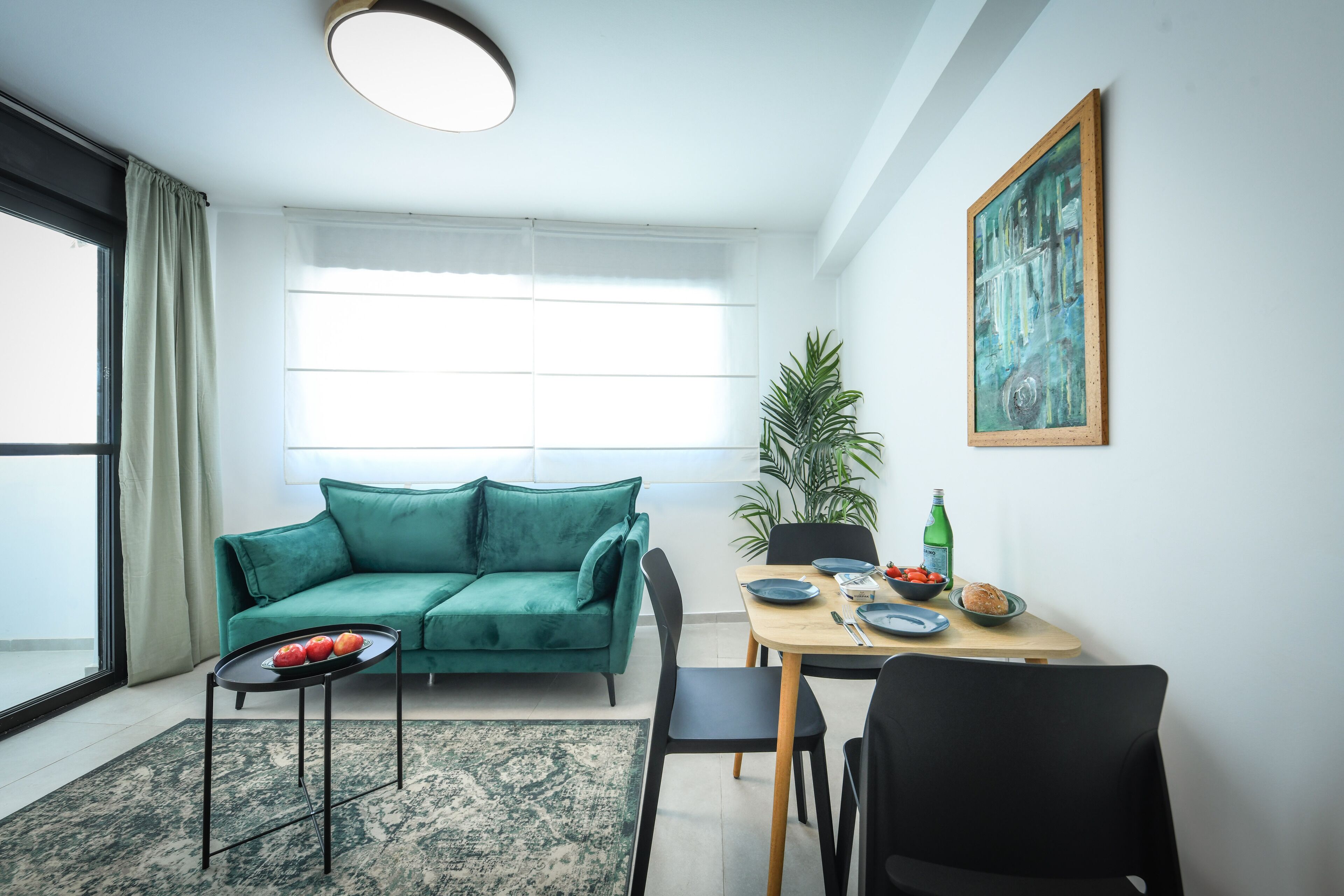 Una sala moderna con un sofá verde, una mesa de comedor con sillas, una mesita con fruta y un ventanal con cortinas.
