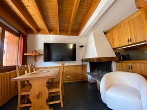 A wooden interior with a dining table, chairs, a flat-screen TV, a fireplace, and a white armchair.