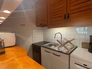A compact kitchen with wooden cabinets, a white tiled backsplash, a stainless steel sink, and a dishwasher.