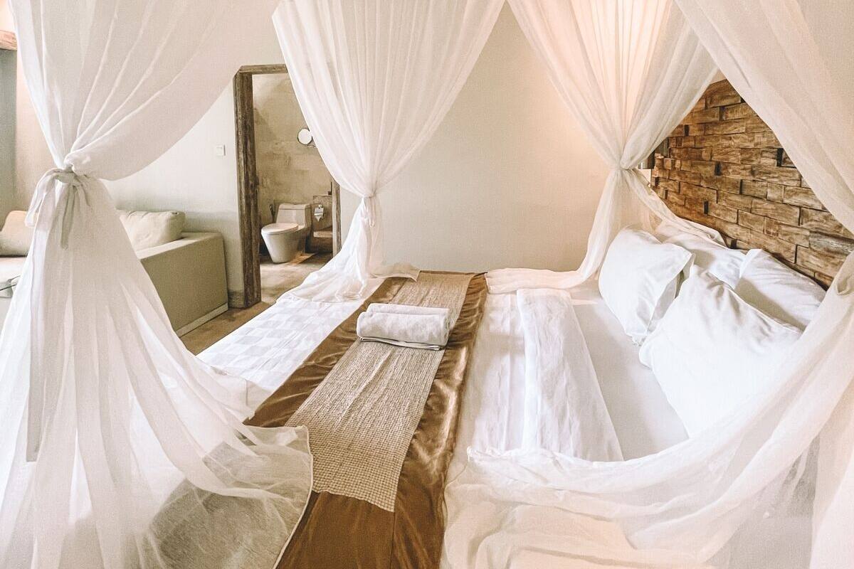 Una cama con dosel sobre cuatro columnas, con un detalle de pared de piedra, cabecera de madera y mesita de noche con lámpara.