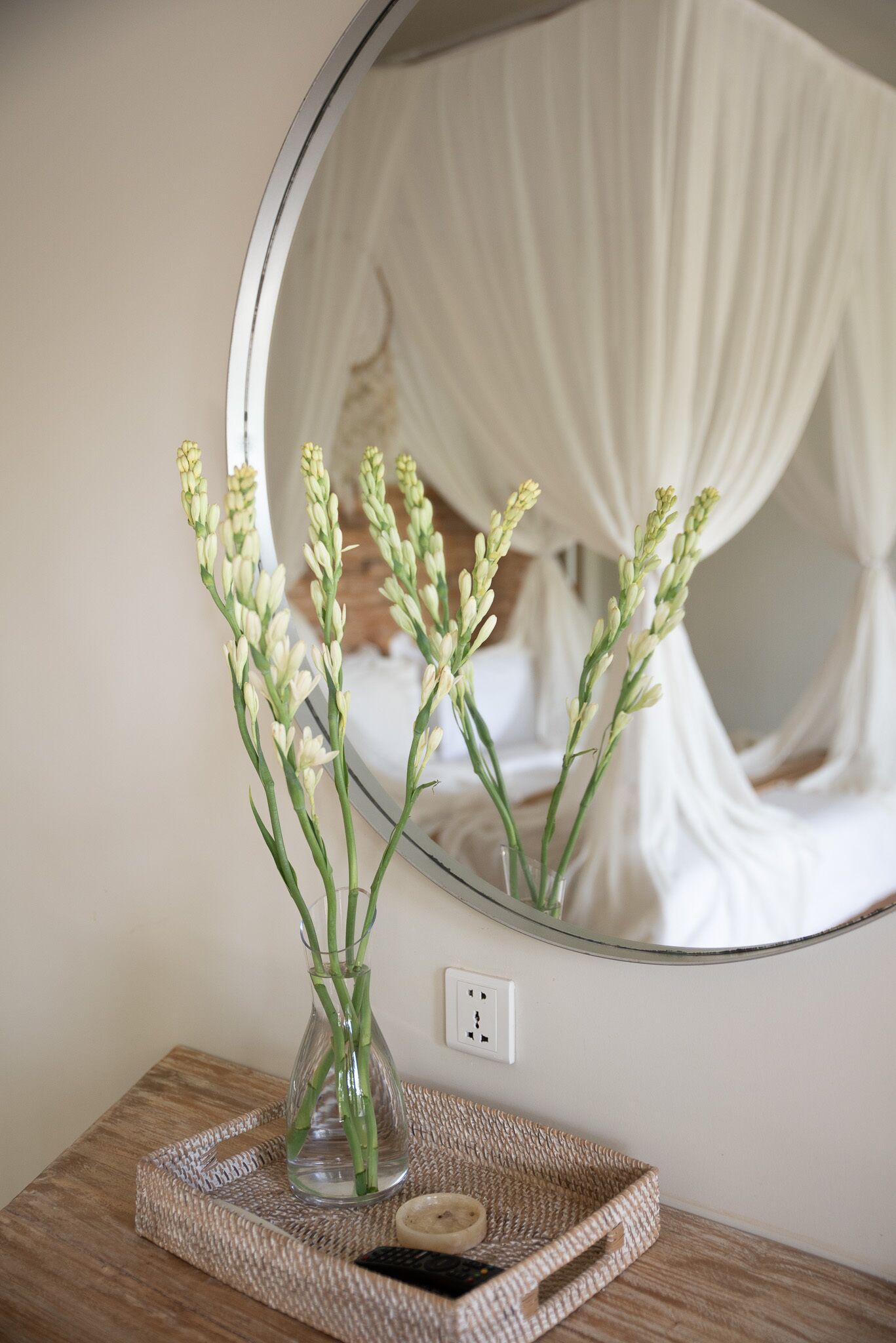 A mirror reflecting a bed with a white canopy, a vase with flowers on a wooden tray, and a power outlet on the wall.