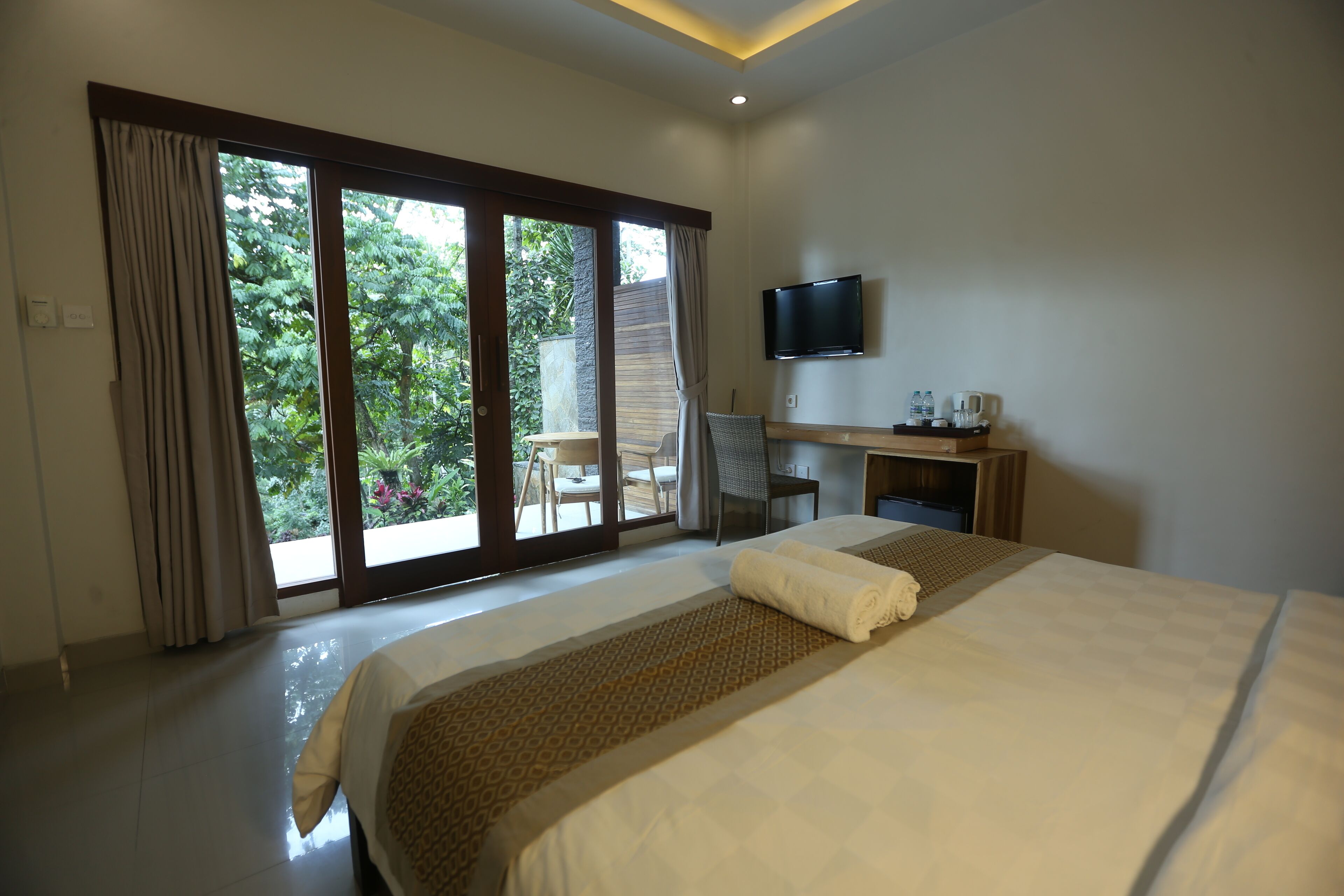 A hotel room with a large bed, a desk with a flat-screen TV, and a view of greenery through the sliding glass door.