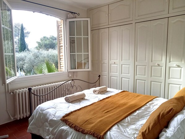 A bedroom with a large window, a radiator, a bed with white bedding and a brown throw, and white wardrobes.