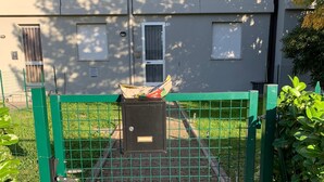 Un espace clos délimité par une clôture en métal, un petit local technique et un chemin pavé menant à un bâtiment doté de fenêtres et d’une porte.