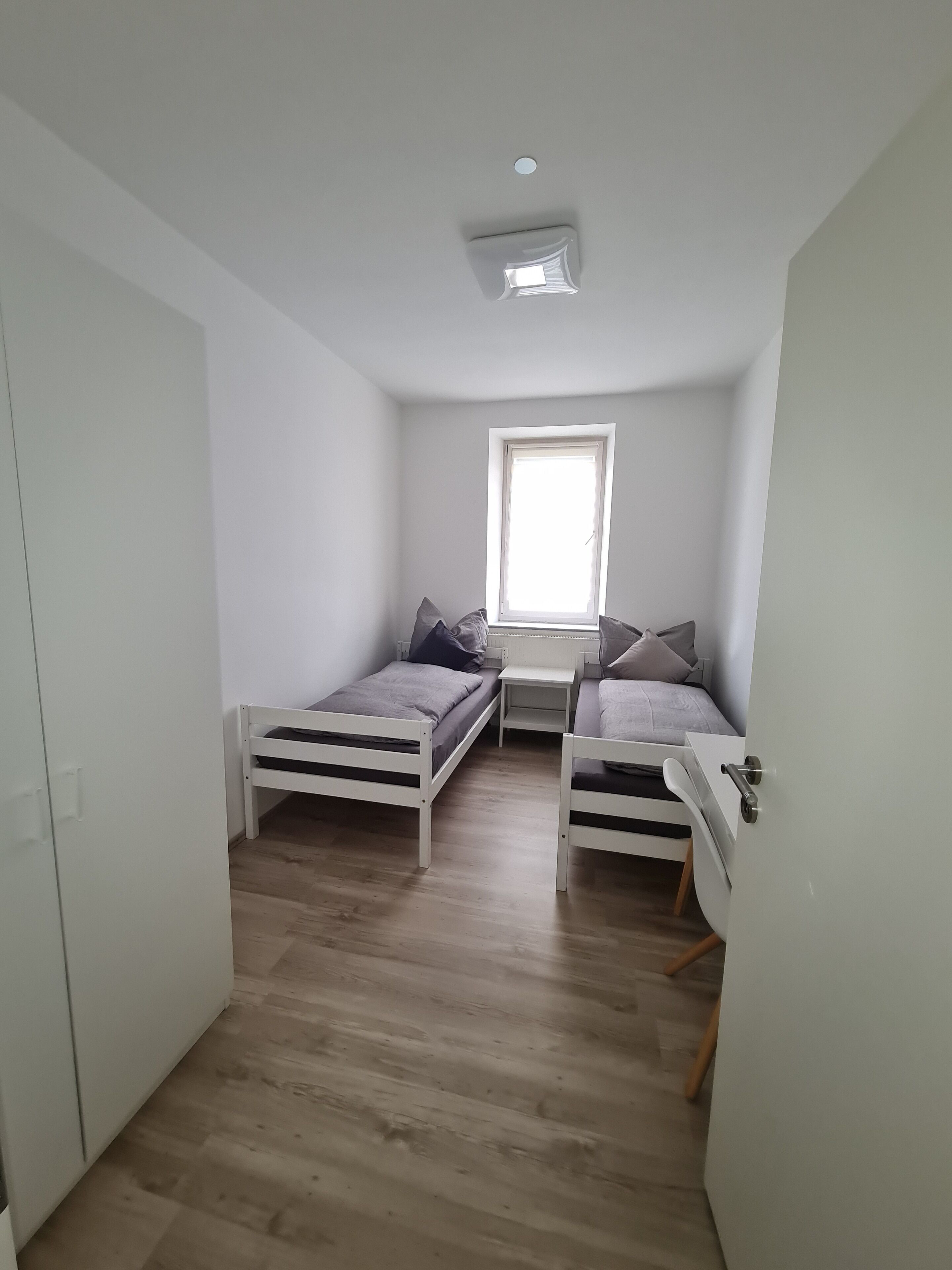 Ein kleiner, gut beleuchteter Raum mit zwei Einzelbetten, einem Fenster und einem weißen Kleiderschrank.